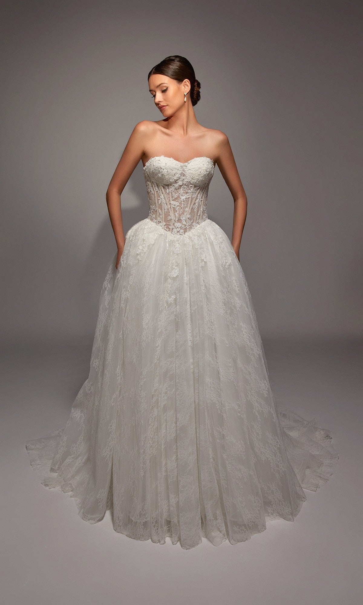 A woman poses in the Alyce 7163, a long white dress with an ivory strapless corset bodice and full lace skirt, hands in pockets, against a plain studio background.